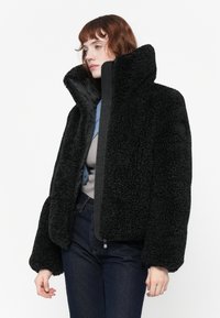 Zwarte fleecejack met hoge kraag, textuur, oversized pasvorm en elastische manchetten, gecombineerd met een grijze ondershirt en blauwe accenten.