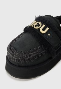 Čierne semišové slip-on topánky s textúrovanými vlnennými akcentmi a zlatým logom na pásku. Majú hrubú čiernu gumovú podrážku a detailné prešívanie.