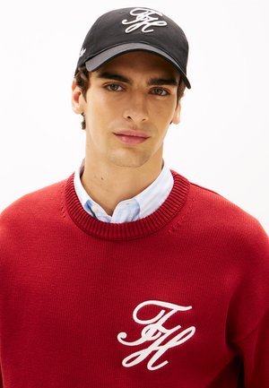Jonge man met een zwarte pet en een rood sweater met witte "FH" initialen, over een lichtblauw gestreept overhemd met kraag, kijkt naar voren.