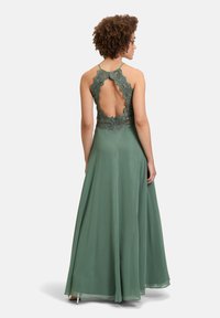 Długa, zwiewna zielona sukienka z koronkową górą, z falbaniastym tyłem i cienkimi ramiączkami, wykonana z gładkiego, teksturowanego materiału.