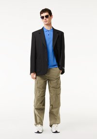 Svart skräddarsydd blazer, blå poloshirt, beige cargobyxor, vita och svarta sneakers, mörka solglasögon. Släta texturer, funktionella fickor.