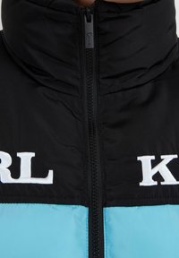 Giacca isolante nera e turchese con colletto alto, chiusura con zip e logo bianco ricamato sul petto. Morbida al tatto, design sportivo.