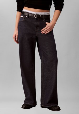Osoba ubrana w ciemne jeansy z szerokimi nogawkami i czarnym paskiem, widoczny biały pas Calvin Klein, czarny krótki top i czarne buty, stojąca na szarym tle.