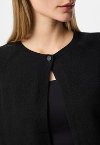 Melna, teksturēta kardigāna ar apaļu kakla izgriezumu, atvērtu priekšpusi un vienu pogu aizdari. Audums izskatās mīksts un ērts.