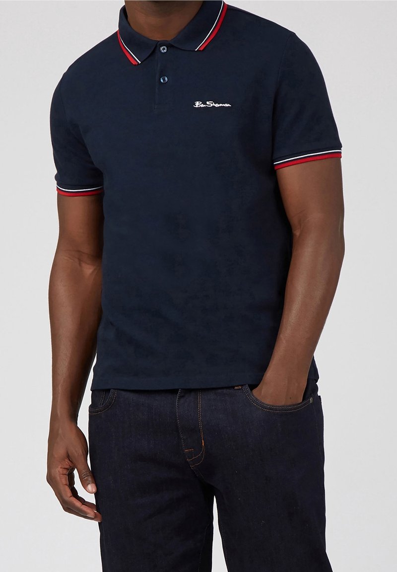 Homme portant un polo bleu marine Ben Sherman avec un col et des bords de manches rayés rouge, blanc et bleu, associé à un jean foncé.