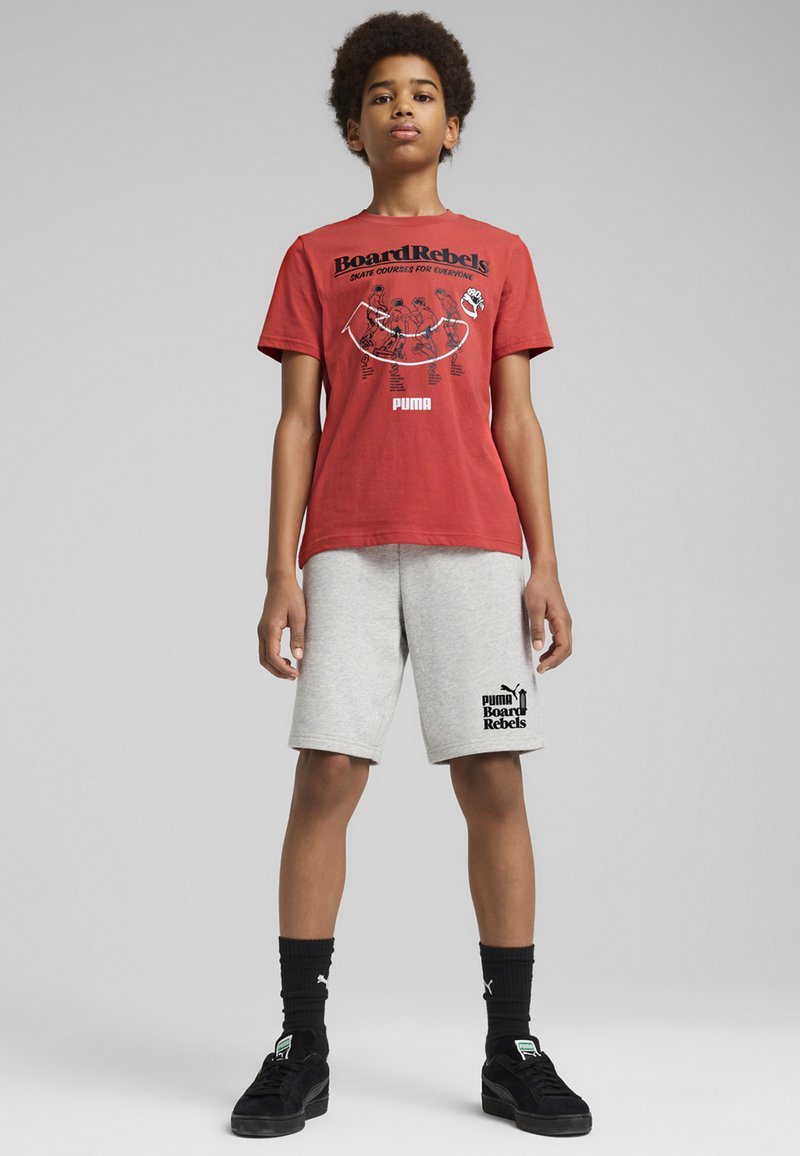 Rdeča bombažna majica s črnimi grafičnimi elementi z napisoma "Board Rebels" in "PUMA"; v kombinaciji s sivo kratkimi hlačami z logotipom; črna obutev.