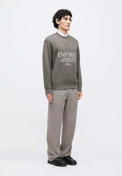 Sivá mikina s bielym nápisom "EMPORIO ARMANI", kombinovaná s svetlosivými nohavicami a čiernymi teniskami. Textúrovaná tkanina s rebrovanými manžetami.