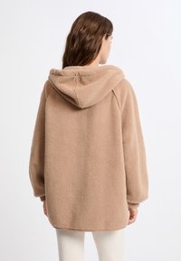 Giacca in pile beige con cappuccio, vestibilità oversize, spalle basse e texture morbida. Presenta un'orlo arrotondato e polsini a coste.