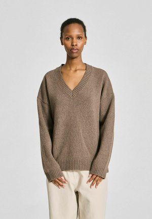 Femme portant un pull en maille marron à col en V et un pantalon beige clair, debout devant un fond clair uni.