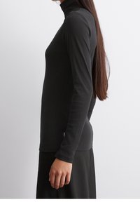 Schwarzes langärmeliges Turtleneck-Oberteil aus Rippstoff, mit einer figurschmeichelnden Silhouette und glatter Textur. Getragen mit einem schwarzen Faltenrock.