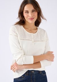 Witte lange mouwen shirt met kantdetails op het voorpaneel, voorzien van een ronde halslijn en knoopsluiting. Combinatie met donkere jeans.