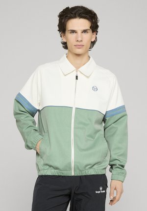 Ung mand med kort krøllet hår iført en lynlåstrøje med hvid top og grøn bund, hænderne i lommerne, neutral baggrund.