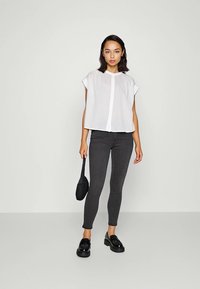 Witte, kortekouter shirt met knoopsluiting, gecombineerd met zwarte skinny jeans en zwarte instappers. Model houdt een zwarte tas vast.
