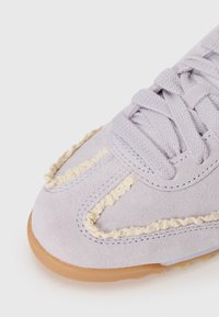 Tenisi din suede violet deschis cu o talpă din cauciuc texturat și accente creamuri răsucite pe lângă cusături. Șireturile și coaserea în nuanțe complementare.