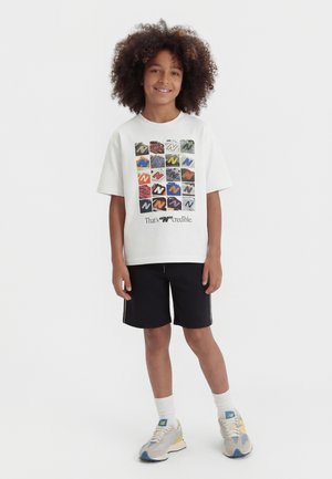 Bambino sorridente con capelli ricci, che indossa una T-shirt bianca con grafica, pantaloncini neri, calzini bianchi e scarpe da ginnastica beige, in piedi su sfondo bianco.