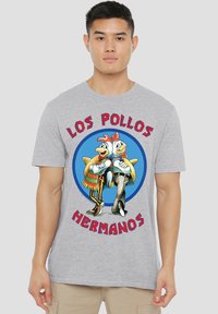 Graues T-Shirt mit cartoonhaften Hühnern in bunten Outfits und dem Text "LOS POLLOS HERMANOS", der einen blauen grafischen Hintergrund umkreist.