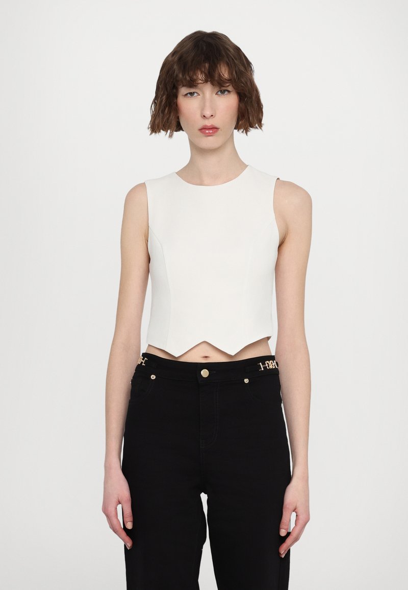 White cropped top with a sleeveless design and a curved, scalloped hem. Paired with high-waisted black jeans featuring gold accents.