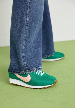 Osoba nosząca zielone i brzoskwiniowe buty Nike z białymi sznurowadłami oraz niebieskie szerokie dżinsy, stojąca na zielonej podłodze.