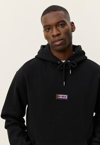 Μαύρο φούτερ με κουκούλα, κατασκευασμένο από βαμβακερό μείγμα υφάσματος, με μπροστινή τσέπη και κουκούλα με κορδόνι. Διαθέτει πολύχρωμο λογότυπο στο στήθος.