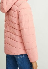 Giacca puffer rosa chiaro con design trapuntato orizzontale, cappuccio e maniche lunghe, abbinata a jeans di denim blu. Texture liscia e stile casual.