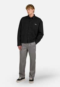 Camisa preta de botão com um bolso frontal e logo branco, combinada com calças cinza claro e ténis pretos com solas brancas.