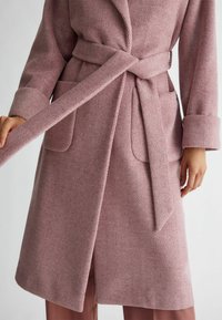 Cappotto con motivo a spina di pesce in tessuto rosa chiaro, caratterizzato da una vita cinturata, tasche ampie e polsini larghi. Texture morbida e design di lunghezza media.