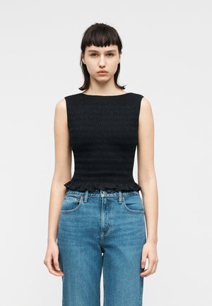 Νεαρή γυναίκα με κοντά σκούρα μαλλιά, που φοράει μαύρο αμάνικο τοπ με ρυτιδώσεις και ψηλόμεσο μπλε τζιν, στέκεται μπροστά σε απλό φόντο.