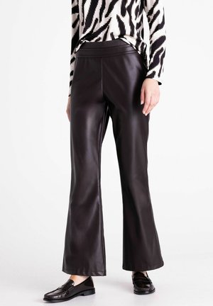 Personne portant un pantalon en cuir noir à jambes larges, des mocassins noirs et un haut à manches longues noir et blanc à motifs, debout sur un fond blanc.