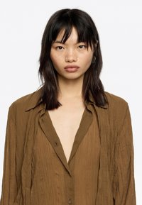 Brown, long-sleeve shirt made of textured fabric, featuring a deep V-neckline and button closures. Loose fit with a slightly wrinkled appearance.