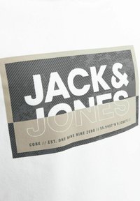 Texte audacieux "JACK &" au-dessus de "JONES" en contour à l'intérieur d'un design rectangulaire noir et beige sur un tissu blanc, avec de petites coordonnées et un texte d'établissement en dessous.