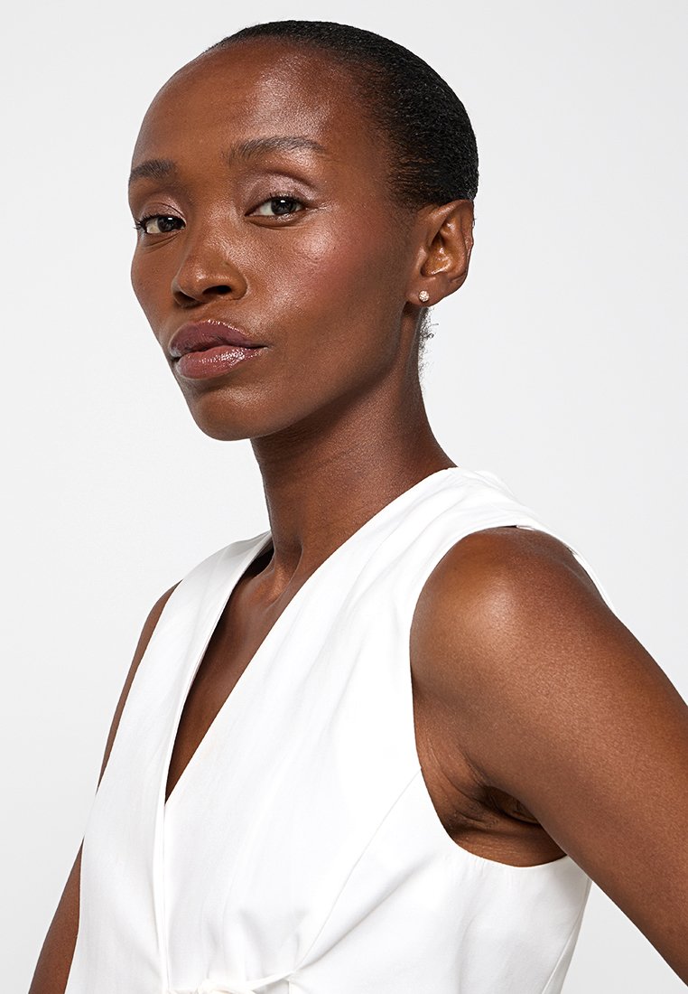 Abito bianco senza maniche con scollatura a V, caratterizzato da una texture liscia e una leggera lucentezza. La modella ha capelli corti e pettinati all'indietro e indossa orecchini di perle.