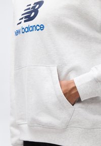 Γκρι ανοιχτό φούτερ New Balance με τσέπη μπροστά, που δείχνει ένα χέρι μισοβυθισμένο στην τσέπη.