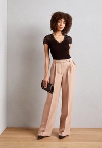 Top negro acanalado con mangas de encaje, combinado con pantalones de talle alto beige. La modelo sostiene un pequeño bolso negro y se encuentra de pie sobre un suelo de madera.