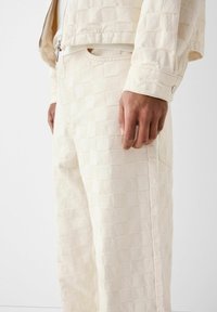 Krämsfärgad bomullsoutfit med ett texturerat rutig mönster, bestående av en kort jacka och avslappnade byxor, samt en synlig sidoficka.