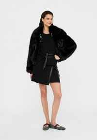 Giacca bomber in pelliccia sintetica nera con maniche in pelle, indossata sopra un top senza maniche e una gonna nera, abbinata a ballerine nere con texture.