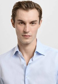 Světlo modra srajca s gumbi na sprednji strani, razprta ovratnica, gladka tekstura in majhni beli gumbi, ki jo nosi model s kratkimi lasmi.