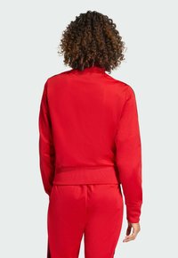 Červená športová bunda s vysokým golierom, rebrovanými manžetami a pásom. Hladká textúra s čiernymi akcentmi na ramenách.