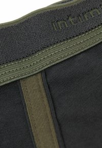 Tejido negro con una superficie texturizada, que presenta una cinturilla elástica de color verde oscuro. Incluye líneas de cosido en contraste y una etiqueta grabada.