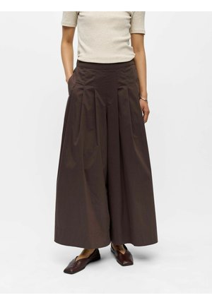 Femme portant un pantalon plissé large marron foncé, un haut beige à manches courtes, des ballerines pointues marron foncé, les mains dans les poches sur un fond blanc.