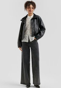 Woman wearing a black leather jacket over a white lace blouse, paired with wide-leg black jeans and black heeled boots, standing with one hand in pocket.