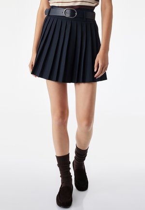 Mujer con una falda negra plisada con un cinturón negro, calcetines marrones y zapatos tipo mocasín de color marrón oscuro, de pie sobre un fondo blanco.