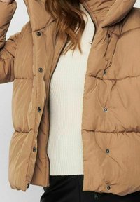 Giacca a vento marrone con cappuccio, caratterizzata da una texture trapuntata, bottoni a pressione e chiusura con zip, indossata sopra una maglia bianca a coste.