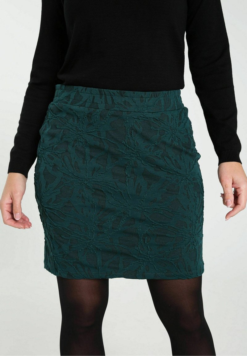 Femme portant une jupe fleurie texturée vert foncé, des collants noirs et un haut noir à manches longues, montrée de la taille au milieu des cuisses.