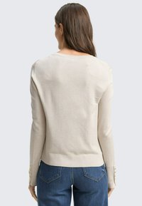 Lichtbeige trui met een aangesloten ontwerp en ribbelzoom, voorzien van drie gouden knoopaccenten op elke manchet. Gecombineerd met blauwe denim jeans.