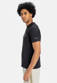 Černé tričko s krátkým rukávem a kulatým výstřihem, které má strukturovanou tkaninu a logo na rukávu. Volný střih, hladká textura.