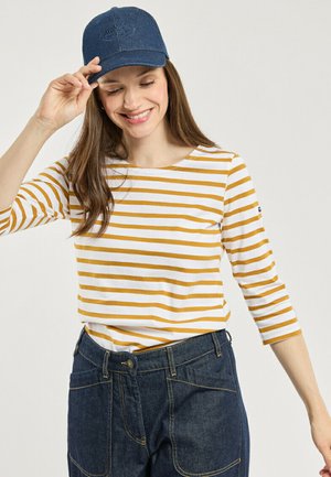 Femme souriante ajustant une casquette bleu marine, portant une chemise rayée blanche et moutarde à manches trois-quarts et un jean en denim foncé taille haute.