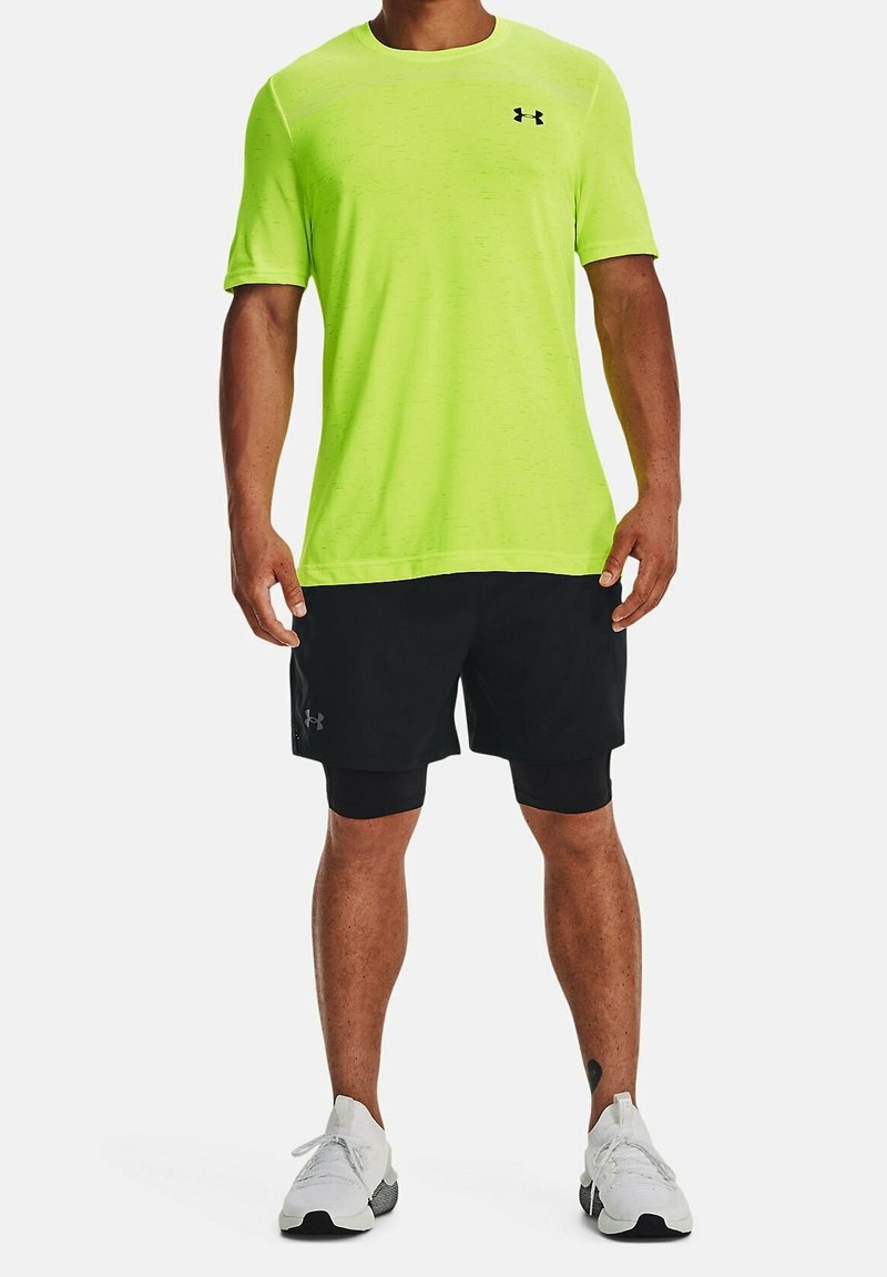 T-shirt de sport vert fluo avec un col rond et des manches courtes, associé à un short noir et des baskets blanches. Logo Under Armour visible.