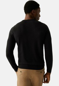 Svart tröja med långa ärmar, ribbad nederkant och ärmslut, tillverkad av ett mjukt tyg. Bärs ihop med ljusa byxor med bakfickor med knappar.