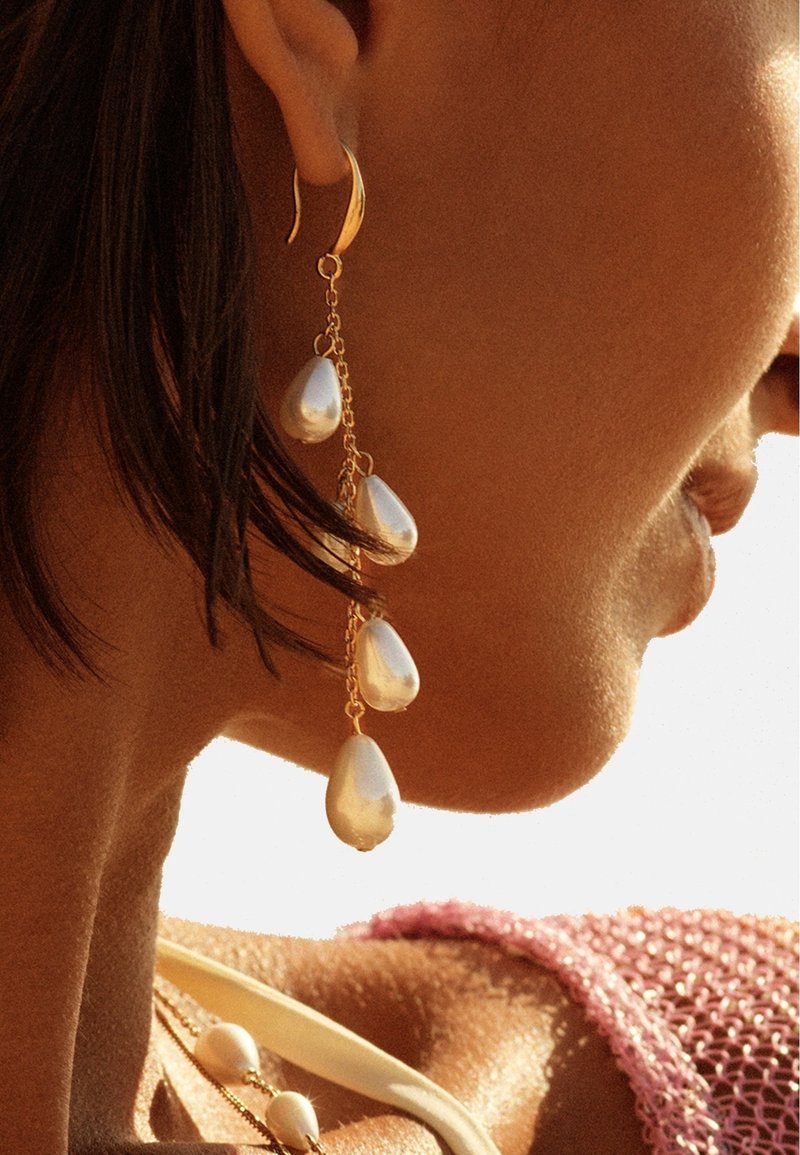 Gros plan sur l'oreille d'une femme portant une longue boucle d'oreille en or avec quatre grosses perles en forme de goutte.