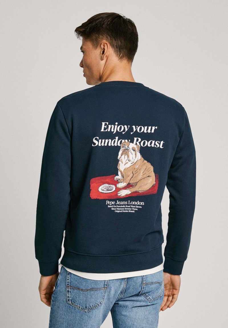 Felpa blu navale con una grafica di un bulldog su un tappeto rosso con una ciotola, testo "Goditi il tuo arrosto domenicale" e dettagli del logo. Materiale in cotone.